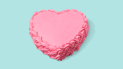 Pink heart-shaped cake with white frosting and pink piping. It's placed against a blue background on a gray surface.